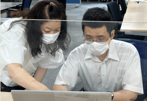 オフィスで透明なパーティション越しに、スタッフがメンバーにノートパソコンで作業内容を説明・指導している様子